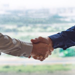 Closeup of two businessmen shaking hands with blurry city view in background. People are seen partly.